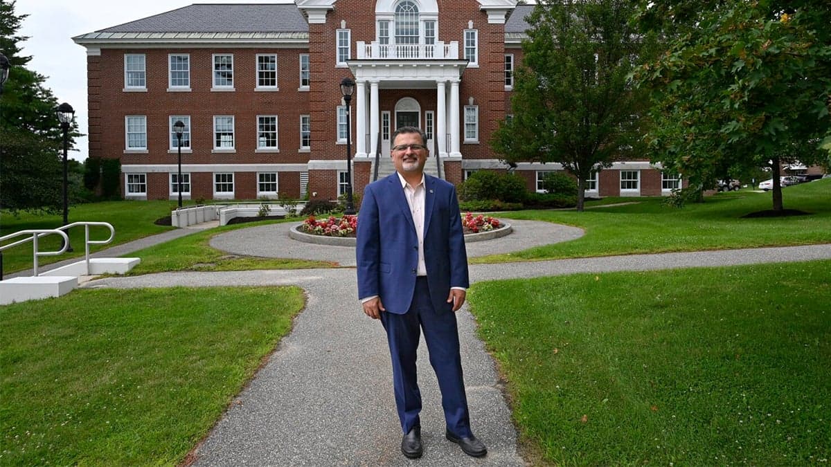 Dr. Melik Peter Khoury, President and CEO of Unity Environmental University, stands smiling in a blue suit on the pathway leading to a traditional brick campus building with white columns and colonial architecture. The three-story administrative building is surrounded by manicured lawns and trees, representing the university's blend of traditional campus aesthetics with innovative educational approaches.